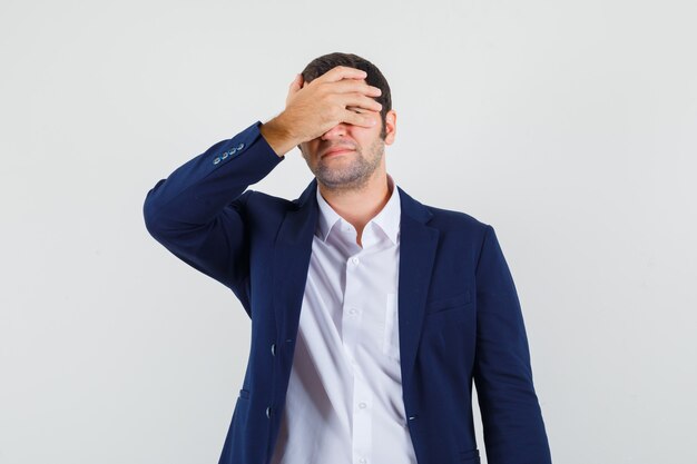 Young male covering eyes with hand in shirt and jacket and looking embarrassed