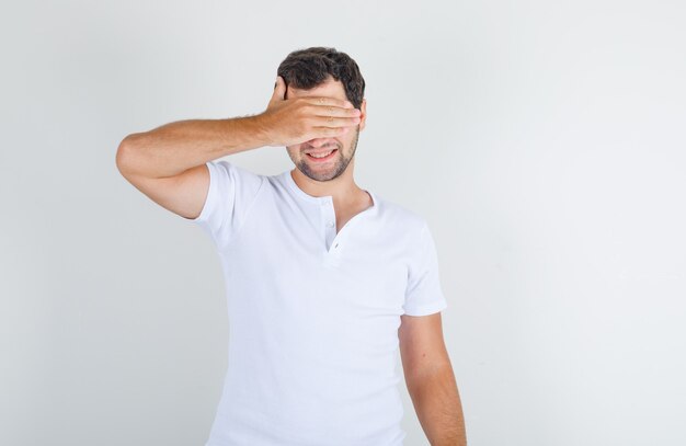 Young male covering eyes with hand in white t-shirt and looking joyful