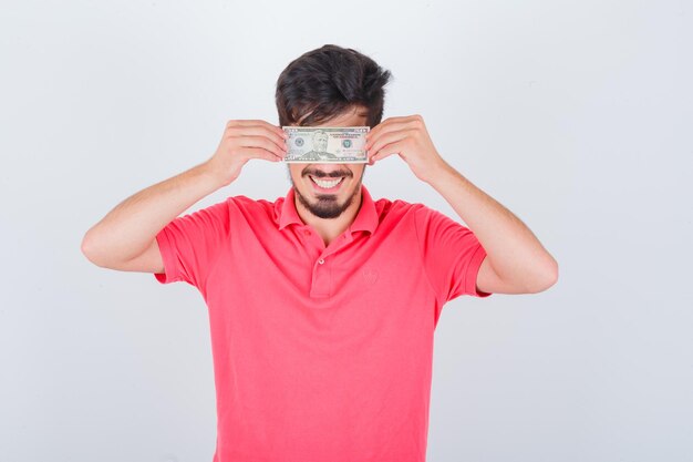 Young male covering eyes with money in t-shirt and looking joyful. front view.