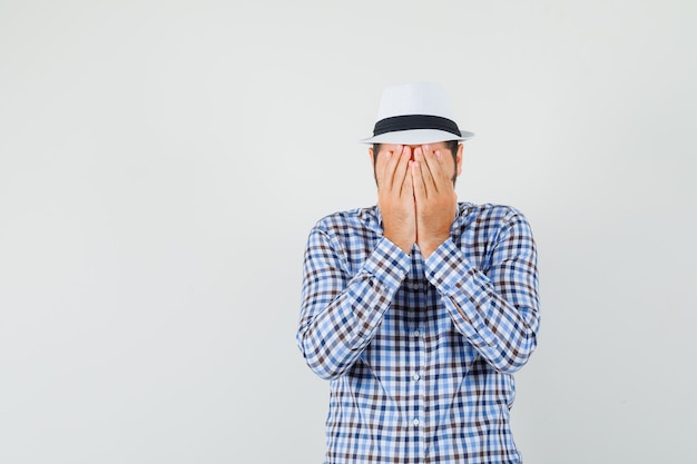 Giovane maschio che copre il viso con le mani in camicia a quadri, cappello e sguardo spaventato, vista frontale.