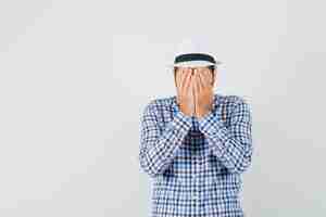 Free photo young male covering face with hands in checked shirt, hat and looking frightened , front view.