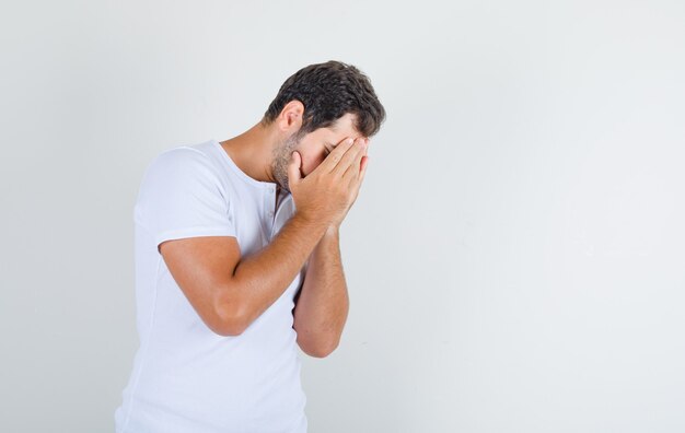 Young male covering his face with hands in white t-shirt and looking sorry