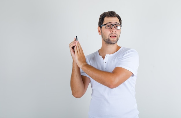 Young male covering microphone on smartphone in white t-shirt, glasses and looking excited