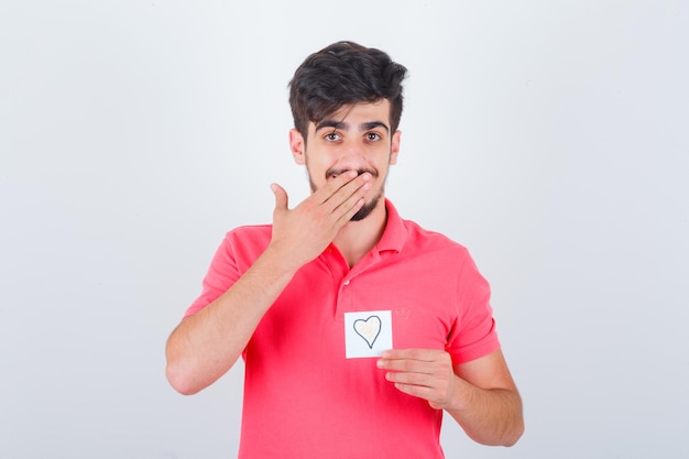 Young male covering mouth with hand in t-shirt and looking pretty , front view.