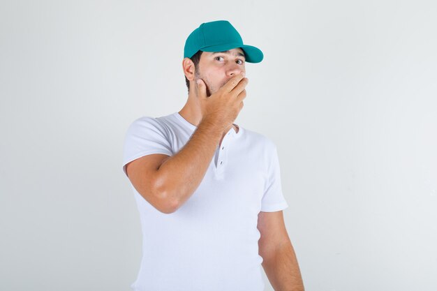 Young male covering mouth with hand in white t-shirt with cap and looking shocked