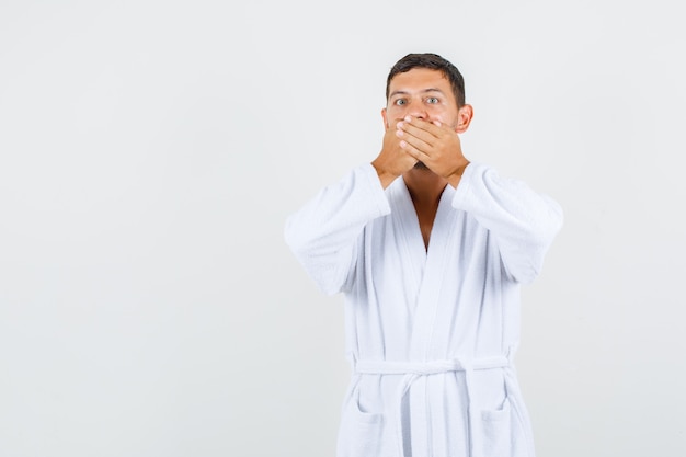 Young male covering mouth with hands in white bathrobe and looking surprised. front view.