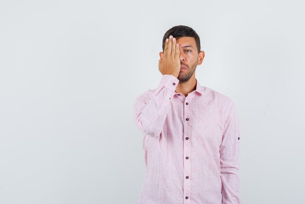 Young male covering one eye with hand in pink shirt , front view.