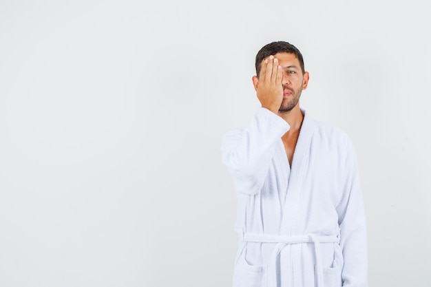 Young male covering one eye with hand in white bathrobe front view.