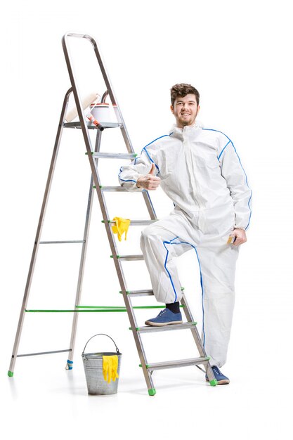 Young male decorator painting with a paint roller and a ladder isolated on white wall.