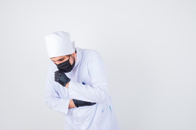 Young male doctor coughing while standing in white uniform and looking unwell. front view.