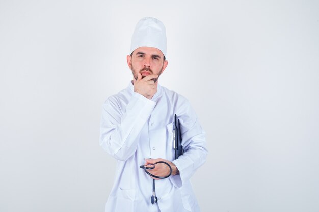 Young male doctor holding clipboard, stethoscope, keeping hand on chin in white uniform and looking thoughtful , front view.