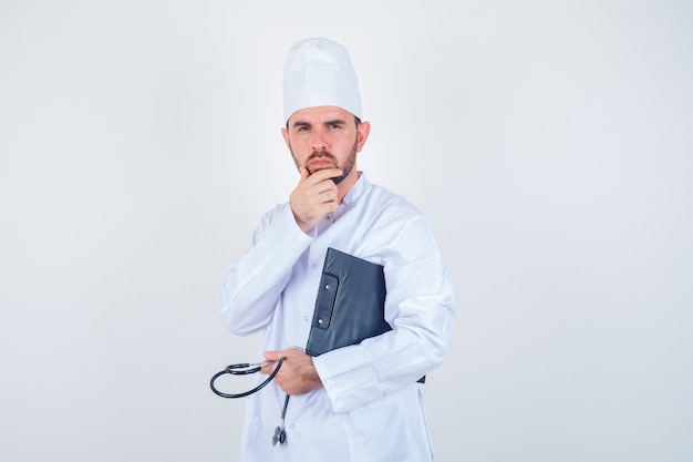 Young male doctor holding clipboard, stethoscope, keeping hand on chin in white uniform and looking thoughtful. front view.