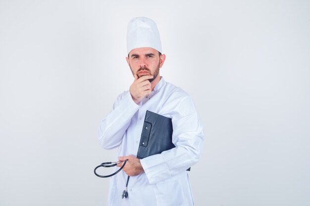 Молодой мужчина-врач, держащий доску сзажимом для бумаги, стетоскоп, держа руку на подбородке в белой форме и выглядящий вдумчивым. передний план.