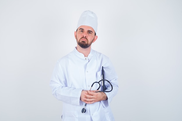 Young male doctor holding clipboard and stethoscope in white uniform and looking careful , front view.