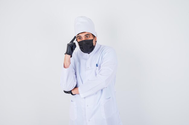 Young male doctor pointing head in white uniform and looking confident , front view.