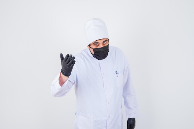 Young male doctor raising hand in questioning pose in white uniform and looking serious. front view.