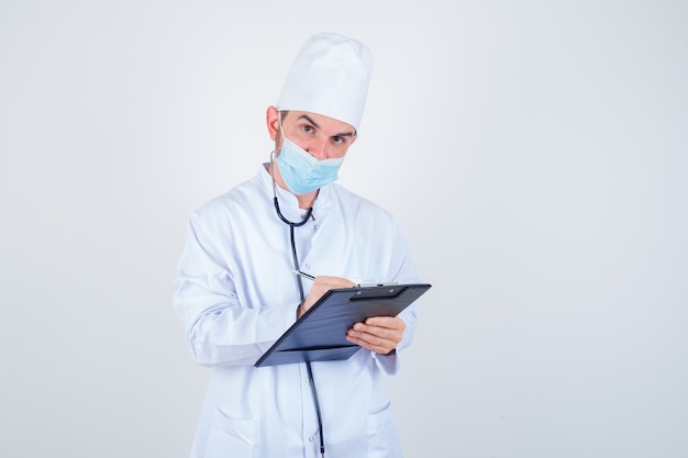 Young male doctor taking notes on clipboard in white uniform, mask and looking sensible , front view.