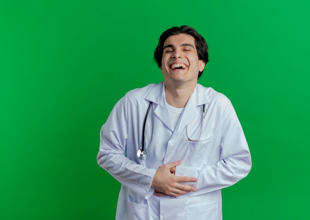 Young male doctor wearing medical robe and stethoscope holding belly and laughing with closed eyes isolated on wall with copy space