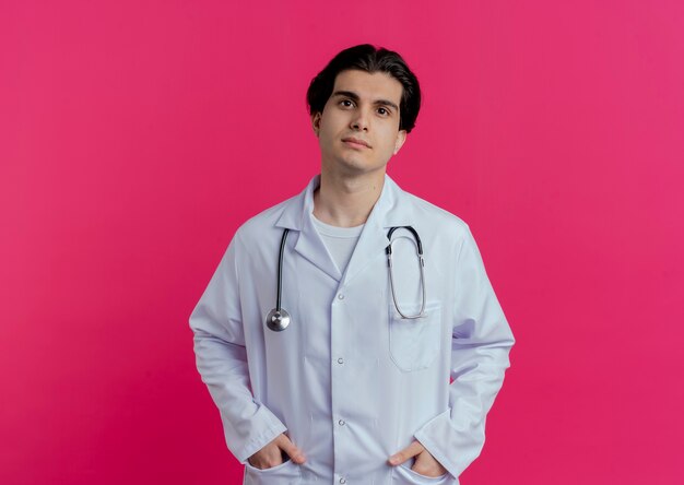 Young male doctor wearing medical robe and stethoscope  putting hands in pockets isolated on pink wall with copy space