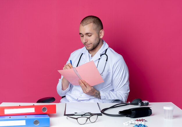 Young male doctor wearing medical robe and stethoscope sitting at desk with work tools holding and looking at note pad and writing something on it with pen isolated on pink background