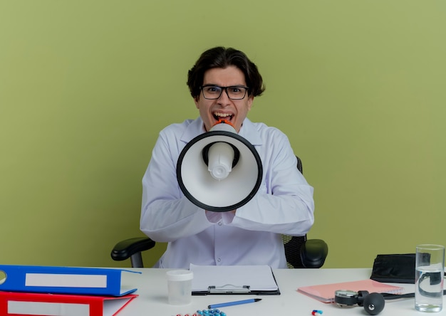 Foto gratuita giovane maschio medico indossa abito medico e stetoscopio con gli occhiali seduto alla scrivania con strumenti medici cercando di parlare da altoparlante isolato