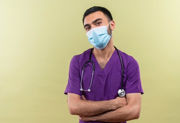 孤立した緑の壁に手を交差させる紫色の外科医の服と聴診器の医療マスクを身に着けている若い男性医師