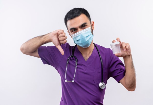 young male doctor wearing purple surgeon clothing and stethoscope medical mask holding empty can his thumb down on isolated white wall