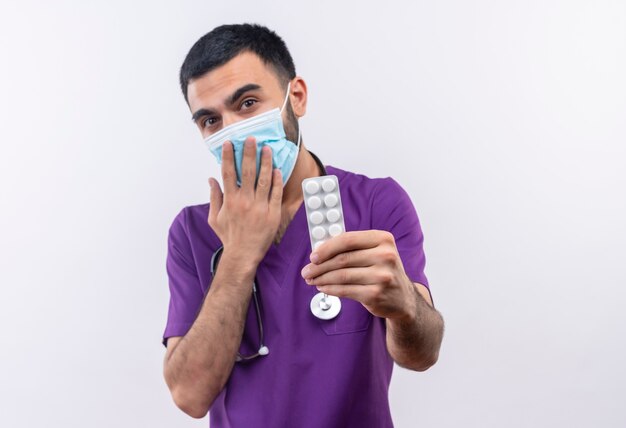 young male doctor wearing purple surgeon clothing and stethoscope medical mask holding out pills to camera covered mouth with hand on isolated white wall