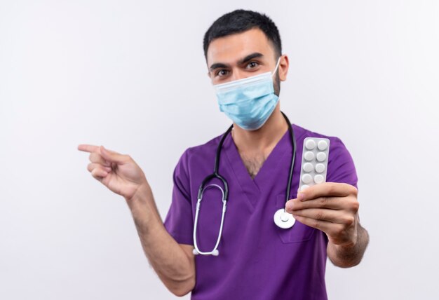 young male doctor wearing purple surgeon clothing and stethoscope medical mask holding out pills to camera points to side on isolated white wall