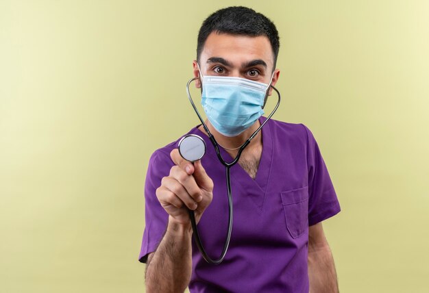 격리 된 녹색 벽에 청진기를 들고 보라색 외과 의사 의류 및 청진 의료 마스크를 착용하는 젊은 남성 의사