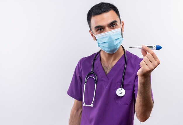 격리 된 흰 벽에 카메라에 온도계를 들고 보라색 외과 의사 의류와 청진 기 의료 마스크를 착용하는 젊은 남성 의사
