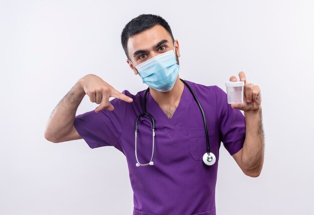 young male doctor wearing purple surgeon clothing and stethoscope medical mask points to empty can in his hand on isolated white wall