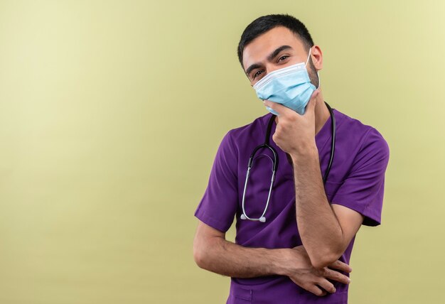 紫色の外科医の服と聴診器の医療マスクを身に着けている若い男性医師は、孤立した緑の壁のあごに手を置きます