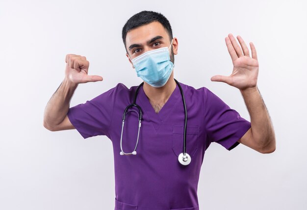 격리 된 흰 벽에 다른 제스처를 보여주는 보라색 외과 의사 의류 및 청진 의료 마스크를 착용하는 젊은 남성 의사