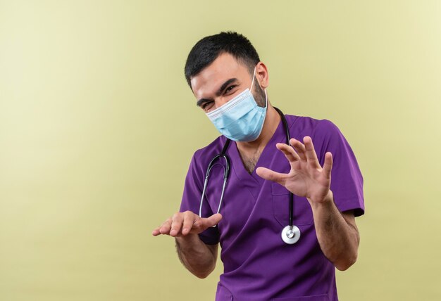격리 된 녹색 벽에 다른 크기를 보여주는 보라색 외과 의사 의류 및 청진 의료 마스크를 착용하는 젊은 남성 의사