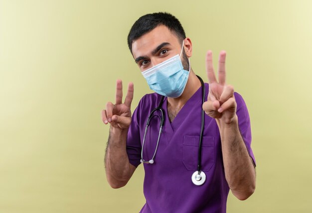 young male doctor wearing purple surgeon clothing and stethoscope medical mask showing peace gesture with both hands on isolated green wall