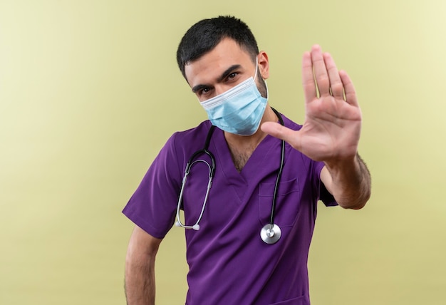 young male doctor wearing purple surgeon clothing and stethoscope medical mask showing stop gesture on isolated green wall