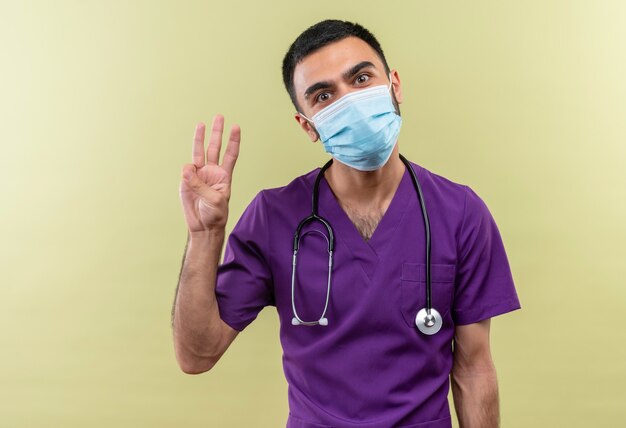 격리 된 녹색 벽에 3을 보여주는 보라색 외과 의사 의류 및 청진 의료 마스크를 착용하는 젊은 남성 의사