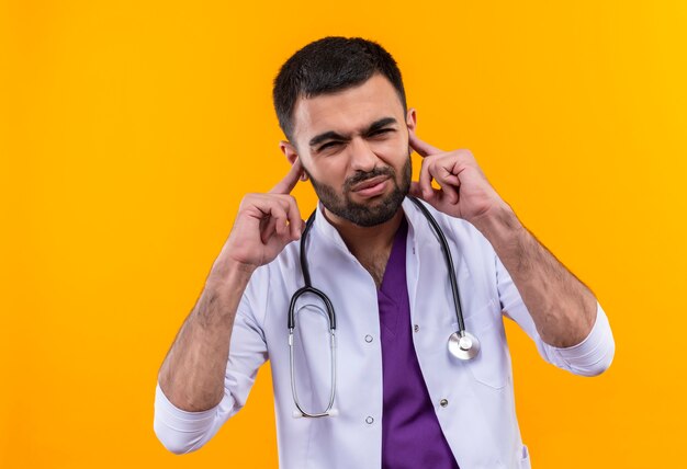 young male doctor wearing stethoscope medical gown closed ears on isolated yellow wall