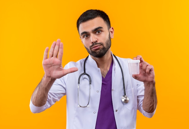 young male doctor wearing stethoscope medical gown holding empty can showing stop gesture on isolated yellow wall