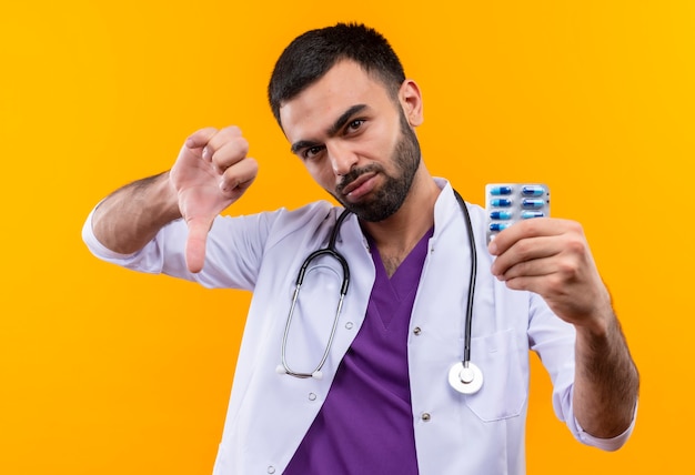 Young male doctor wearing stethoscope medical gown holding pills his thumb down on isolated yellow wall