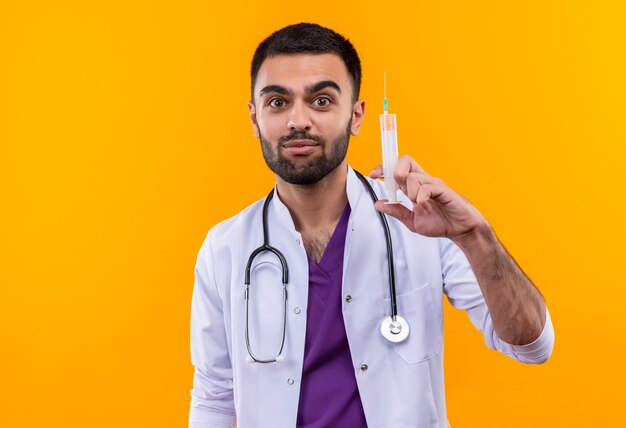 молодой мужчина-врач в медицинском халате со стетоскопом держит шприц на изолированной желтой стене