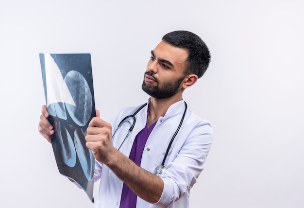 Молодой мужчина-врач в медицинском халате со стетоскопом смотрит на рентгеновский снимок в руке на изолированном белом