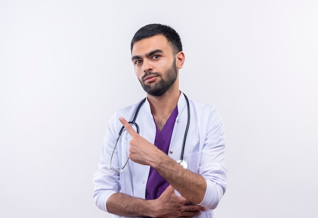 young male doctor wearing stethoscope medical gown points to side on isolated white wall