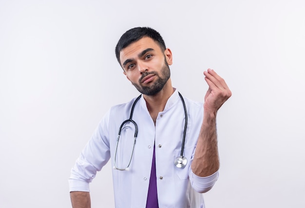 молодой мужчина-врач в медицинском халате со стетоскопом показывает денежный жест на изолированной белой стене