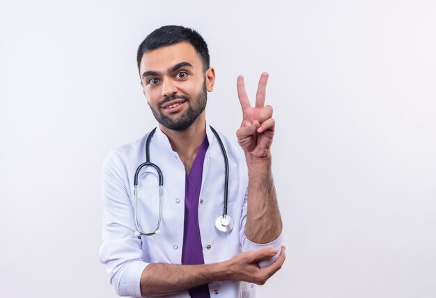 Молодой мужчина-врач в медицинском халате со стетоскопом показывает жест мира на изолированной белой стене