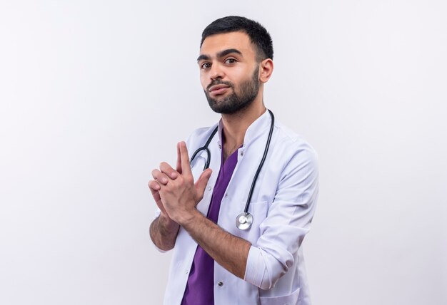Бесплатное фото Молодой мужчина-врач в медицинском халате со стетоскопом показывает жест пистолета на изолированной белой стене