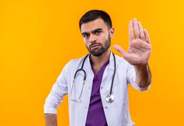 young male doctor wearing stethoscope medical gown showing stop gesture on isolated yellow wall