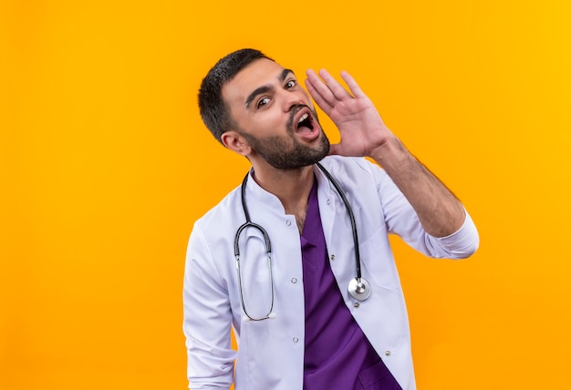 young male doctor wearing stethoscope medical gown whispers on isolated yellow wall