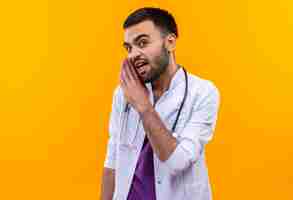 Free photo young male doctor wearing stethoscope medical gown whispers on isolated yellow wall